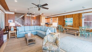 A spacious living area with a light blue sectional sofa, a wooden dining table with chairs, and a staircase with wooden railings. - Gulf Front House in Orange Beach | Direct Beach Access | Pool | Sleeps 24 (Orange Beach)