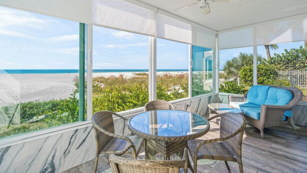 A sunlit room with a glass table and wicker chairs, overlooking a beach and ocean. - Island House Beach Resort 1N (Siesta Key)