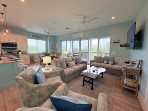 A spacious living room with a kitchen, a ceiling fan, and large windows.