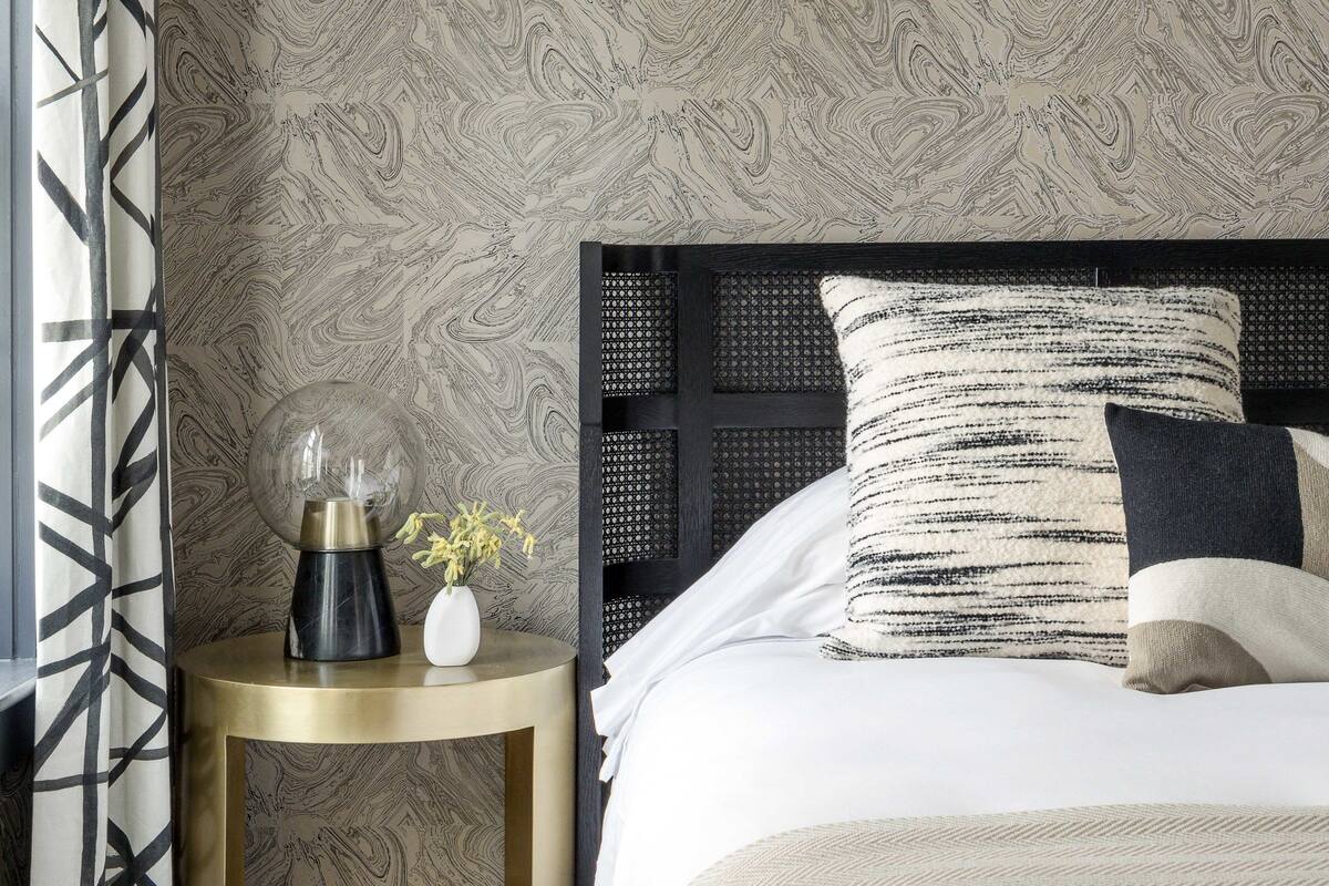 Une chambre à coucher avec un papier peint à motifs, une table de chevet ronde surmontée d’un vase, et un lit agrémenté de plusieurs coussins.