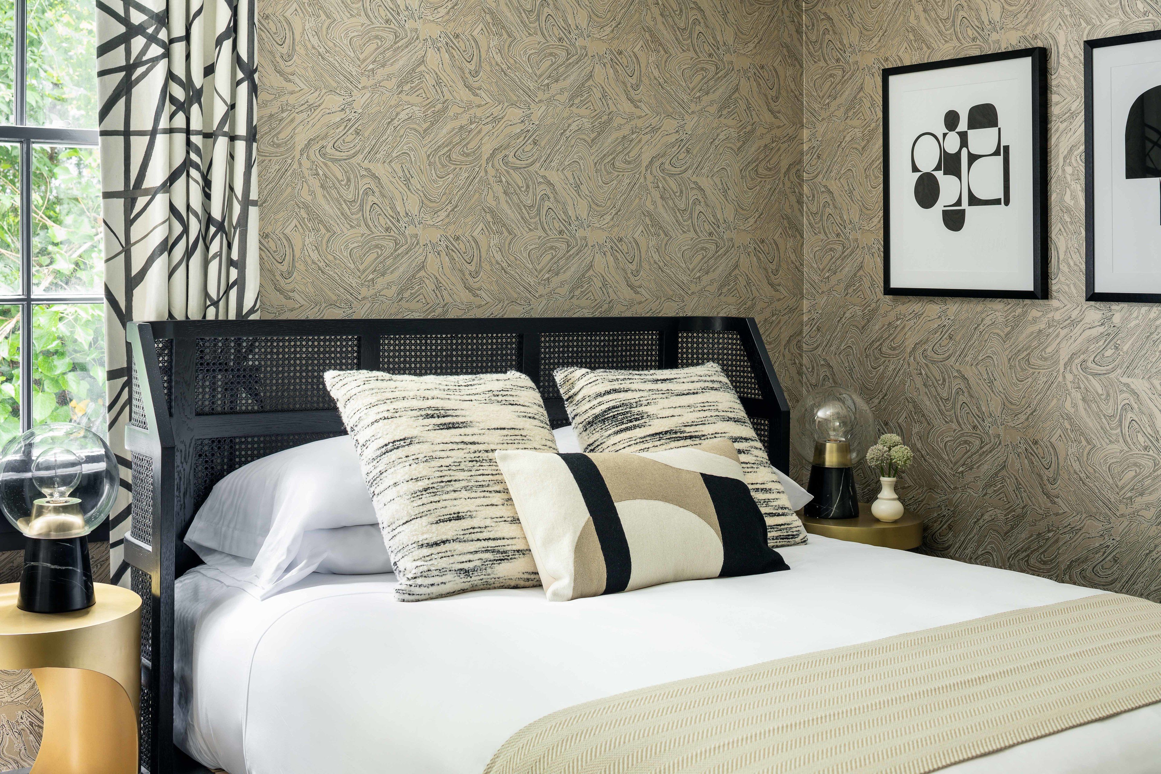 Un letto rifatto con cura, con testiera nera, cuscini bianchi e decorati, e una coperta beige. È presente un comodino con una lampada e un vaso di fiori. La stanza presenta carta da parati decorata e una finestra con tende.