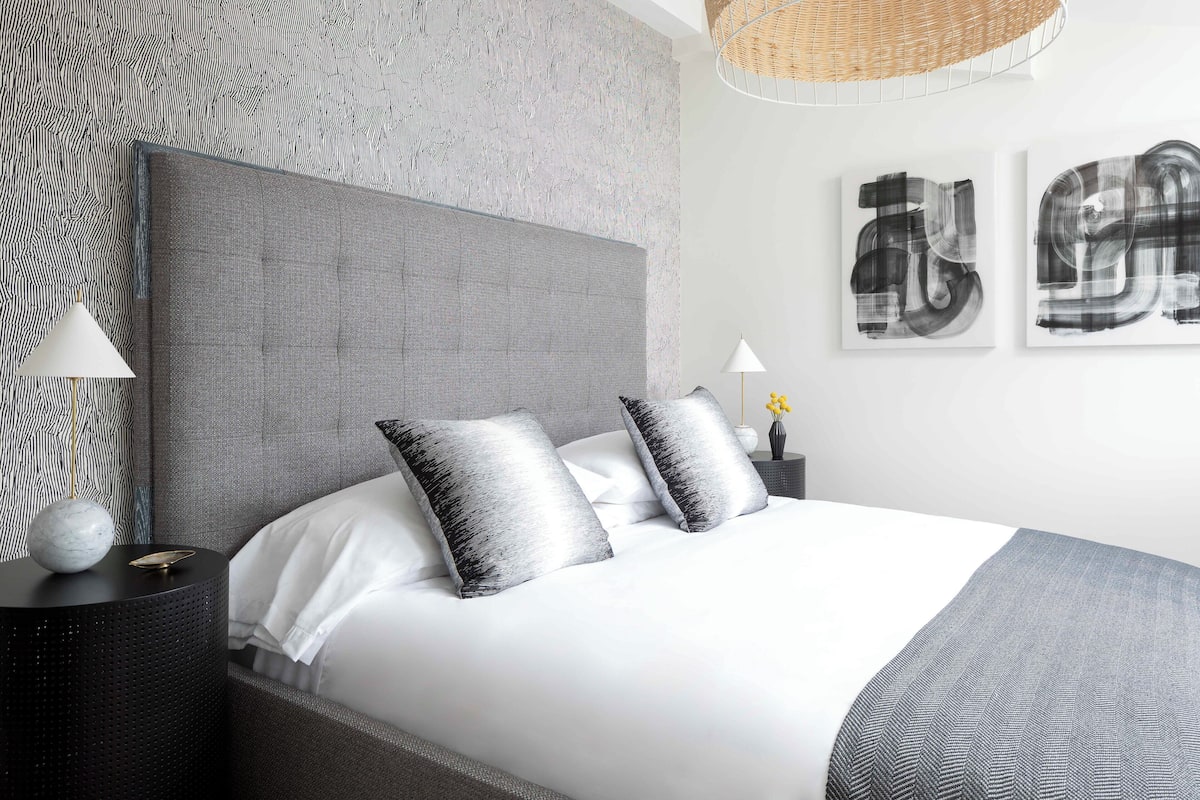 Une chambre moderne avec un grand lit, une tête de lit grise et deux tables de chevet avec des lampes.