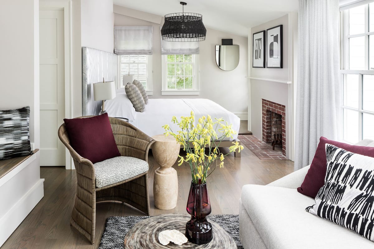 Une chambre à coucher avec un lit, une chaise, un canapé, un vase de fleurs, une cheminée et une fenêtre.