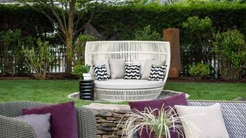 A patio area with outdoor furniture, including a wicker sofa and a cushioned bench, surrounded by greenery and a stone fire pit.