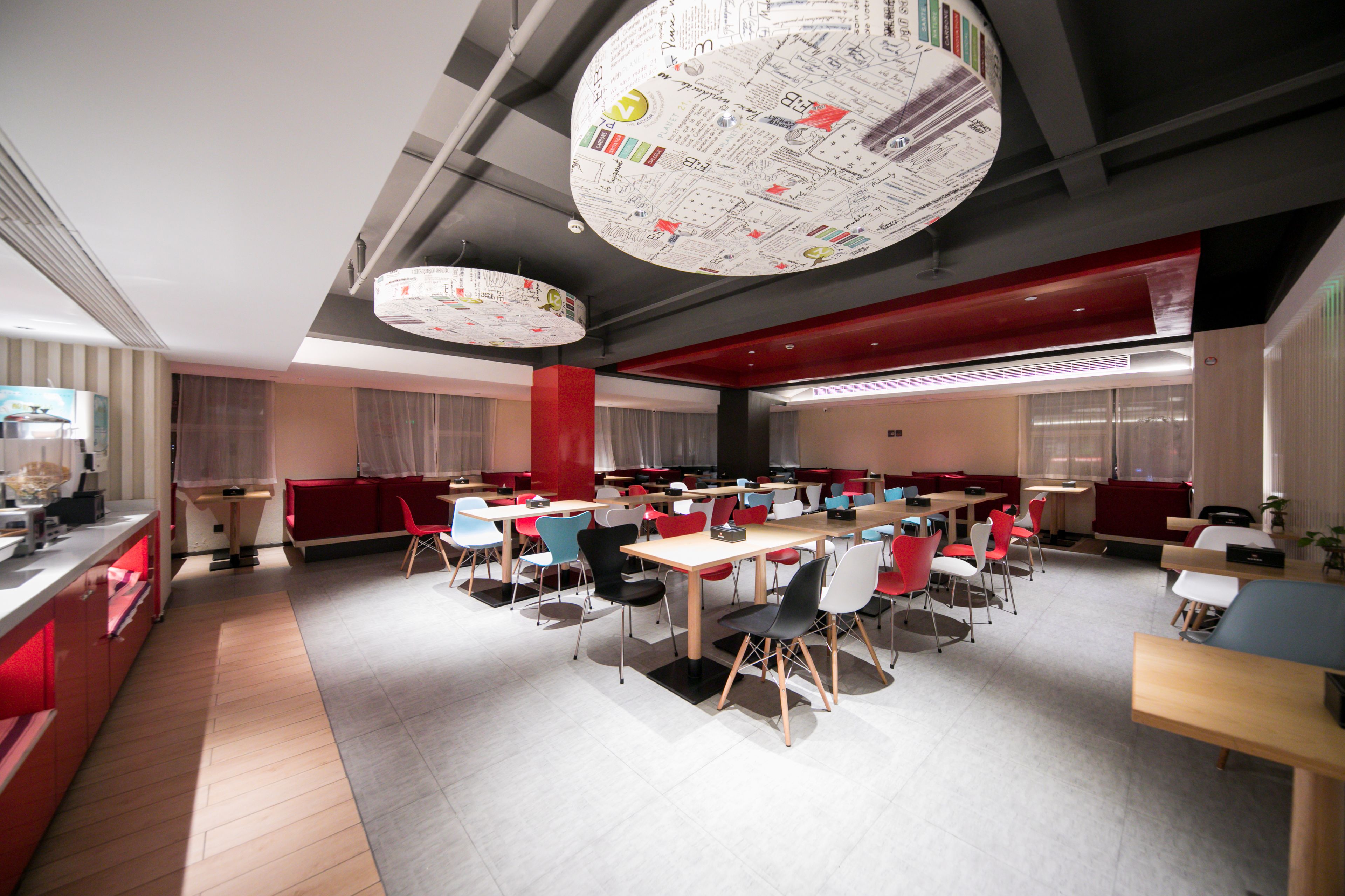 A modern dining area with multiple tables and chairs, a red accent wall, and a ceiling with circular light fixtures.