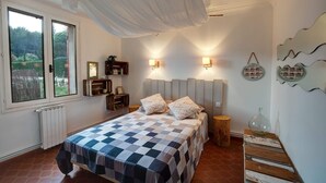 A bedroom with a checkered bedspread, a window with a view of greenery, wall-mounted shelves, and a decorative wall piece. - Vacation home in Luberon in the heart of Provence WIFI Free Fiber (Lauris)