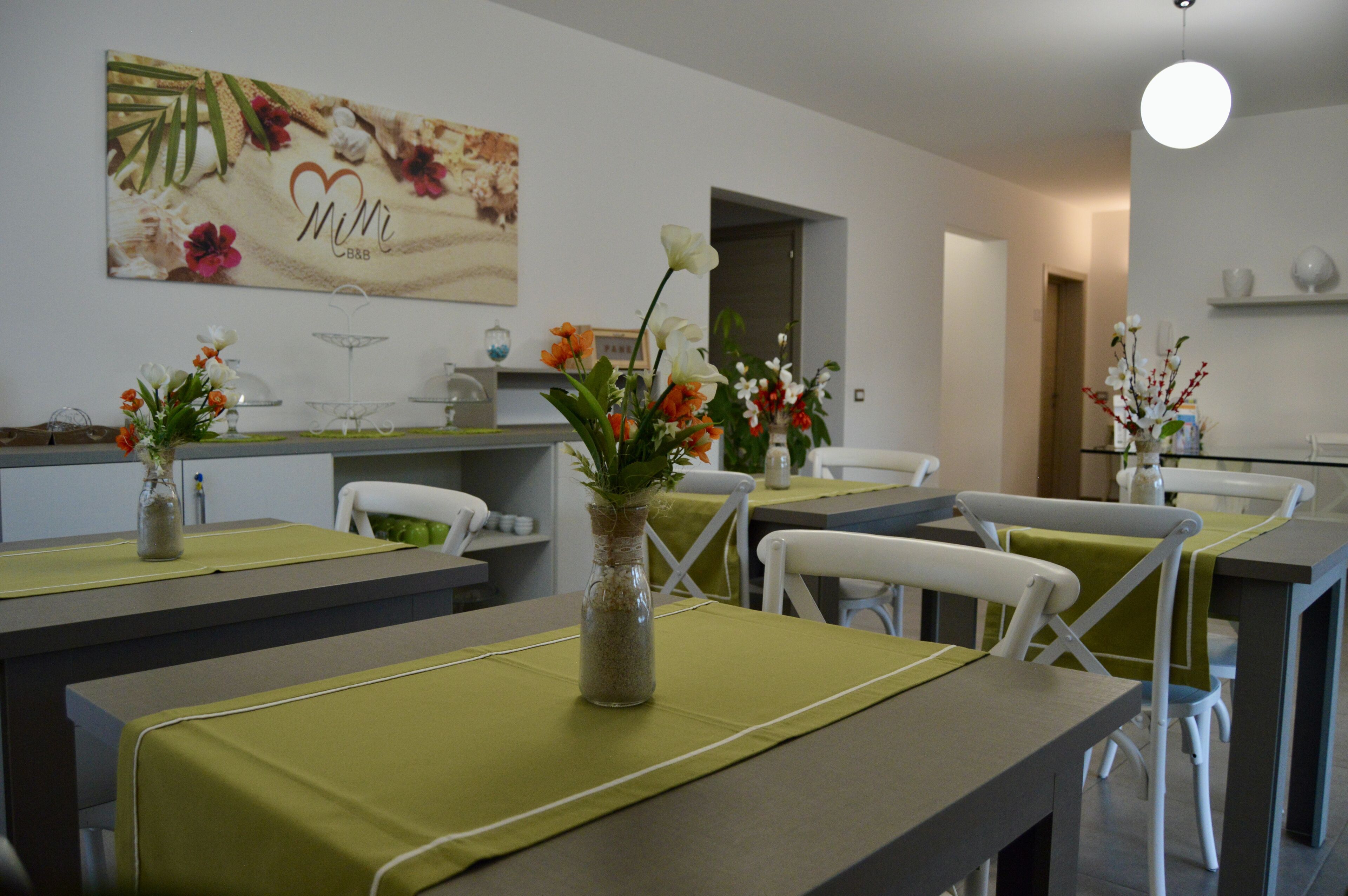 A dining area with tables set for service, a vase with flowers on each table, and a wall with a floral painting.