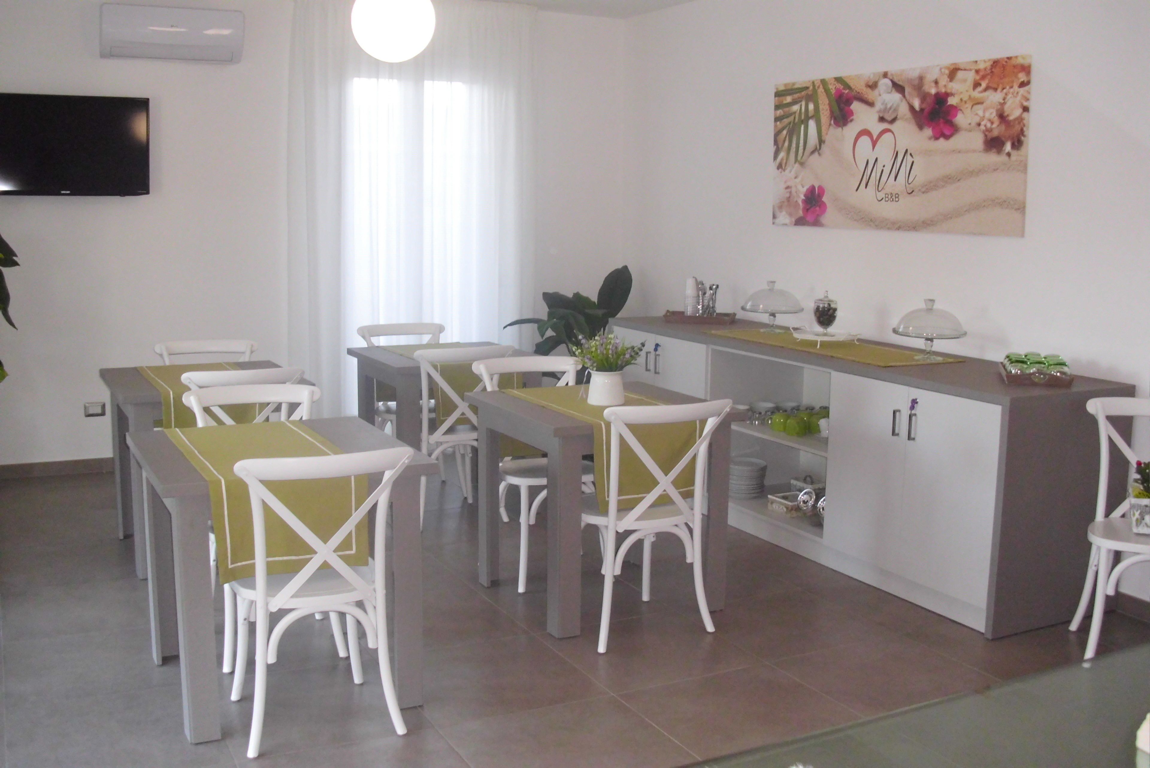 A dining area with white chairs and tables, a kitchen counter with a plant, and a wall-mounted TV.