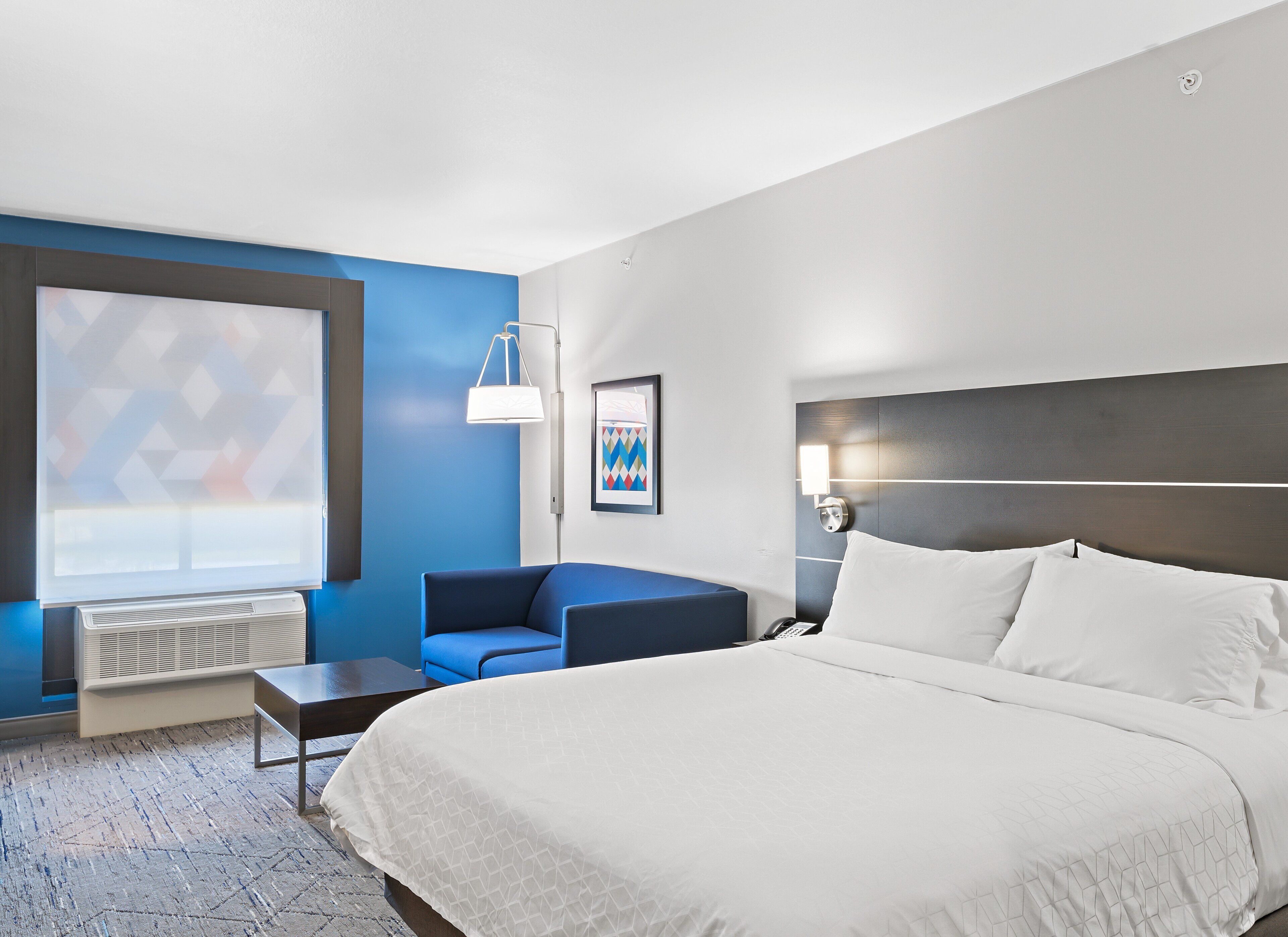 A hotel room with a large bed, a blue armchair, a small table, a desk, a desk lamp, and a window with a geometric view.