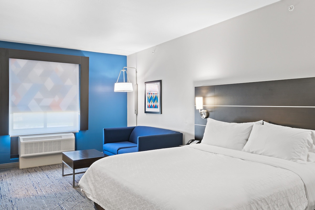 Habitación de hotel con una cama grande, un sillón azul, una mesita, un escritorio, una lámpara de escritorio y una ventana con vistas geométricas.