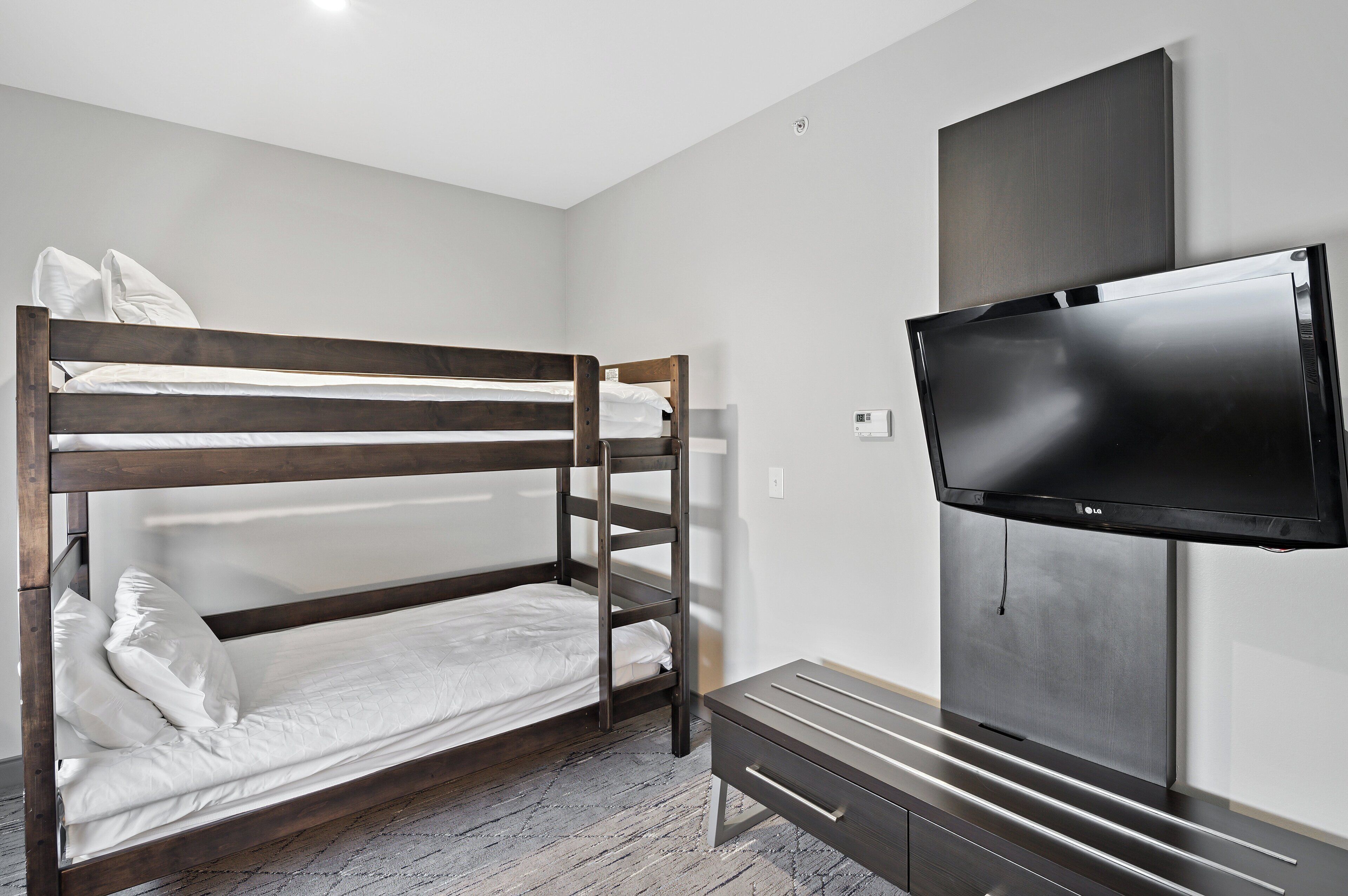 A bunk bed with white bedding, a flat-screen TV mounted on the wall, and a wooden bench.