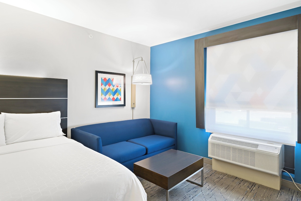 Una habitación de hotel con cama, sofá azul, una mesita, lámpara y un ventanal con persianas.