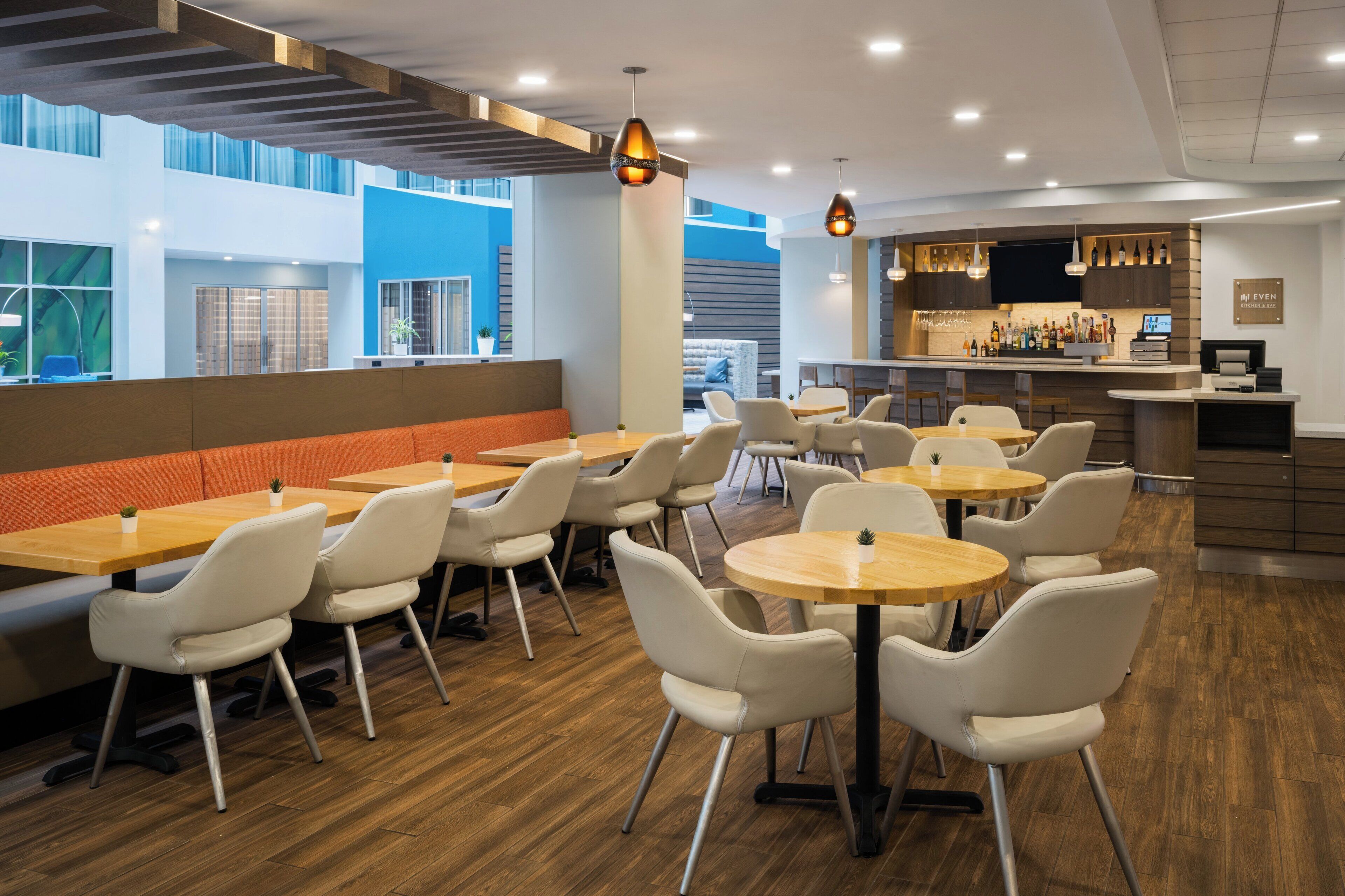 A modern restaurant with wooden tables and white chairs, a bar area, and large windows offering views of the outside.