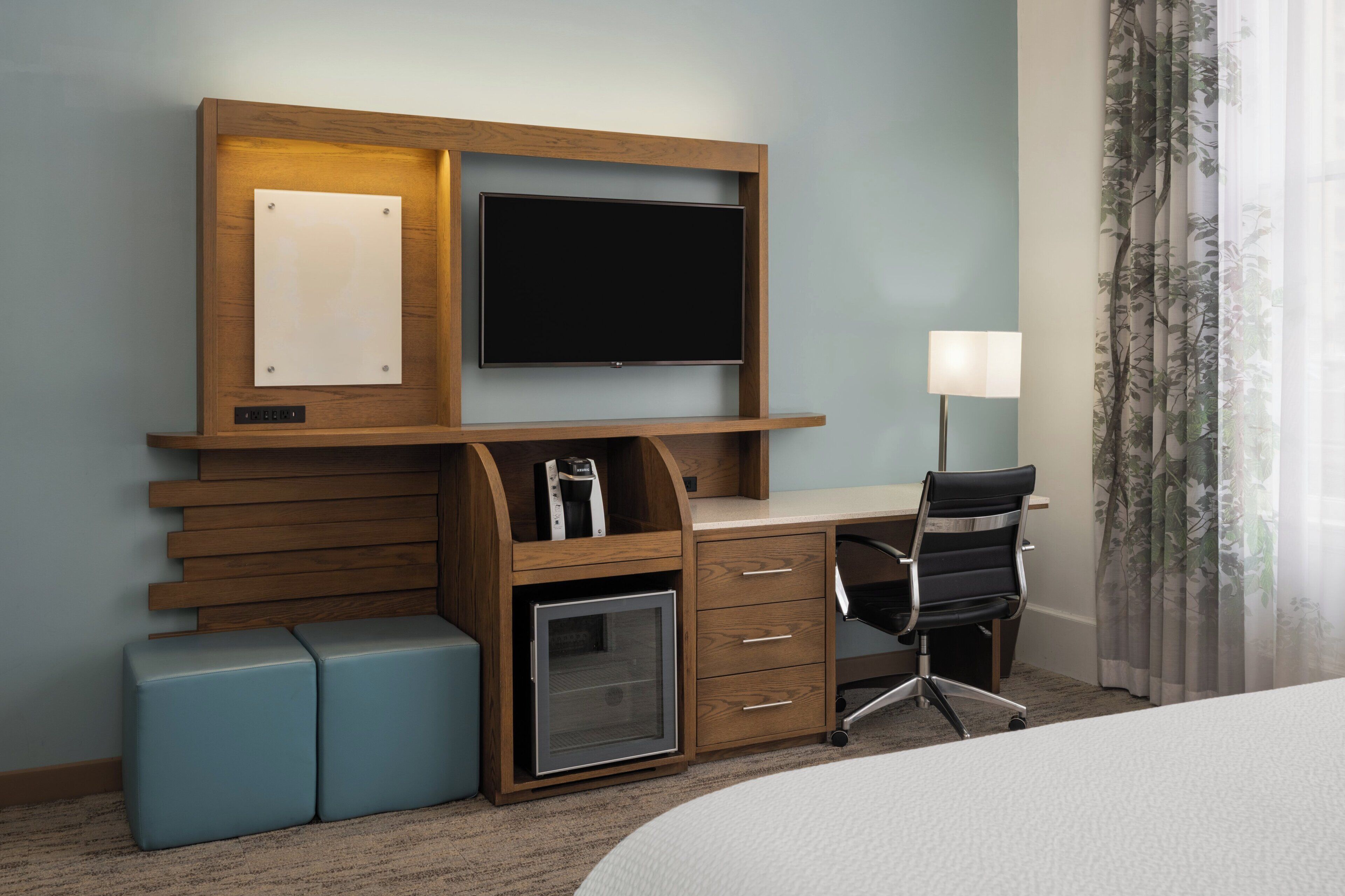 A hotel room with a wooden desk, a flat-screen TV, a small refrigerator, a chair, and a bed.