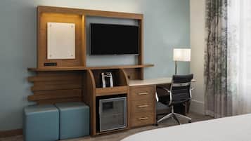 A hotel room with a wooden desk, a flat-screen TV, a small refrigerator, a chair, and a bed.