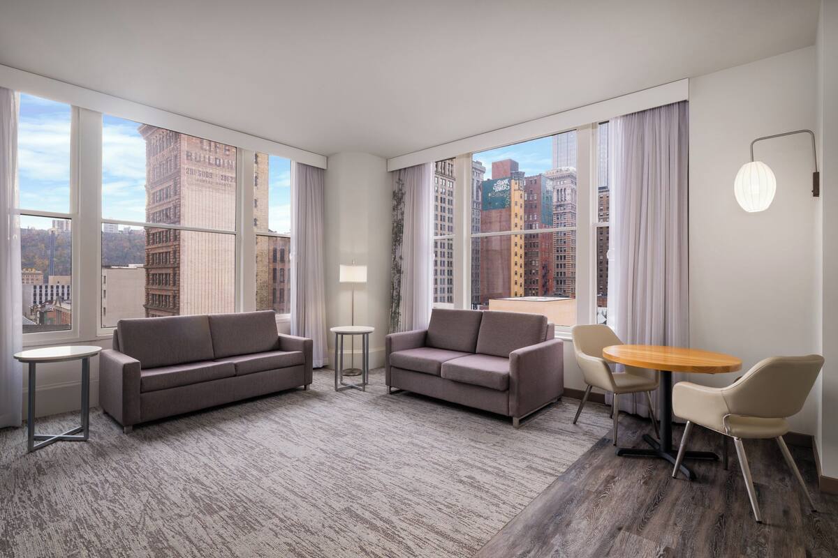 A modern hotel room with a large window offering a city view, a sofa, a small round table, and a lamp.