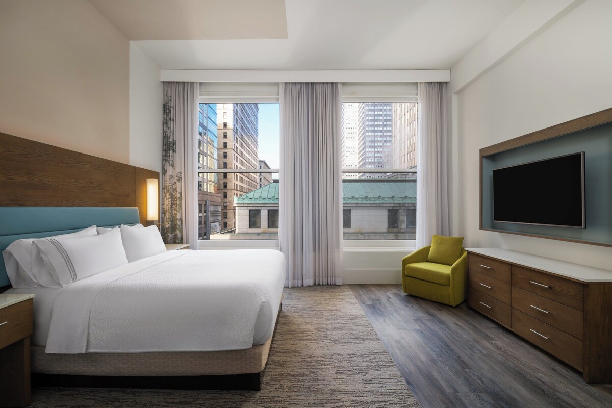 Habitación de hotel moderna con una cama grande, un televisor montado en la pared, un sillón amarillo y vistas a la ciudad a través de amplios ventanales.