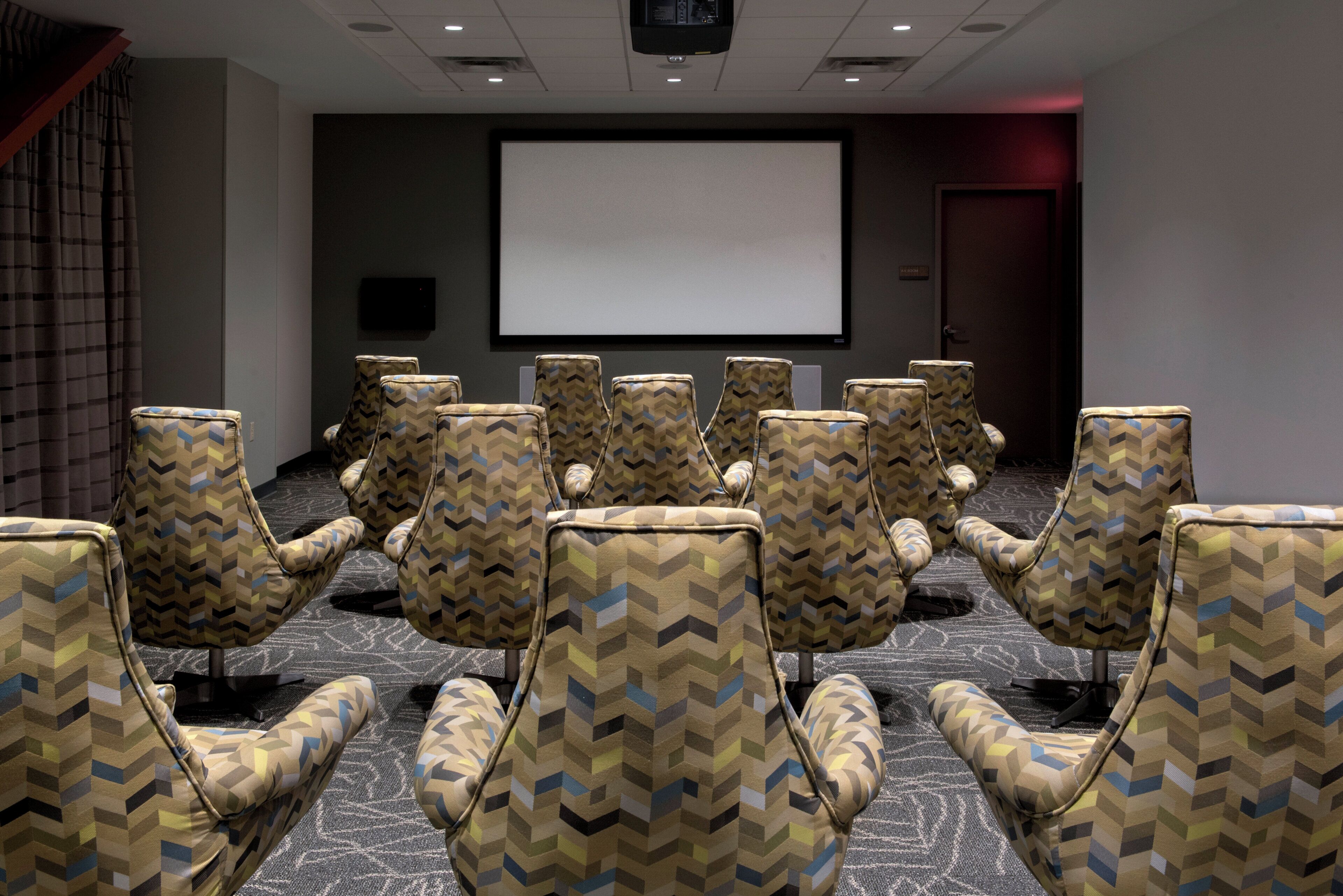 A conference room with patterned chairs arranged in rows, a large screen, and a projector.