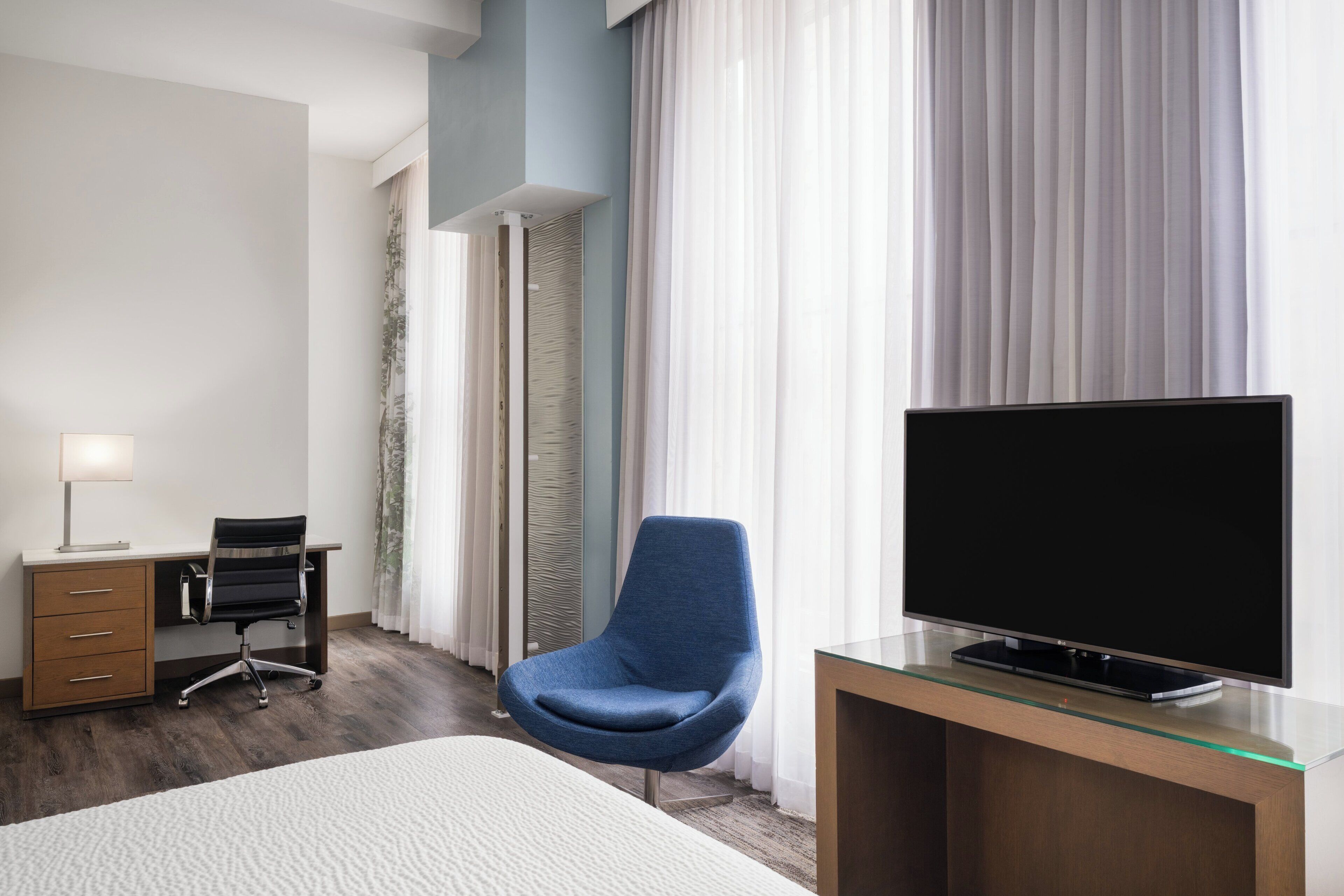 Quarto de hotel com cama, escrivaninha com computador, cadeira, televisão e janela com cortinas.