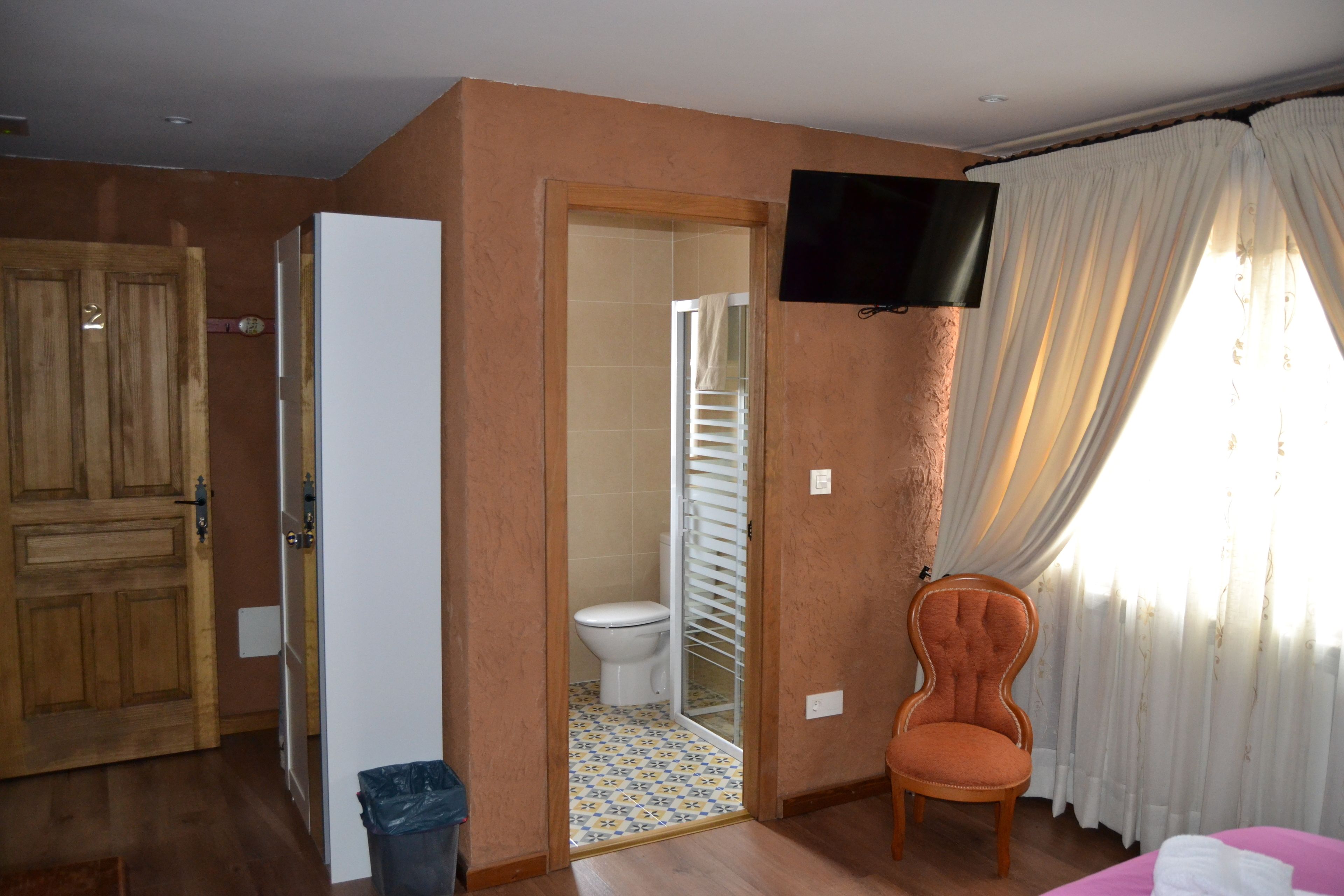A hotel room with a wooden door, a television mounted on the wall, a chair, and a bed.