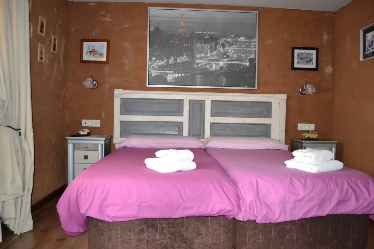 A double bed with pink bedding, two bedside tables with lamps, a framed cityscape picture on the wall, and a curtain on the left.