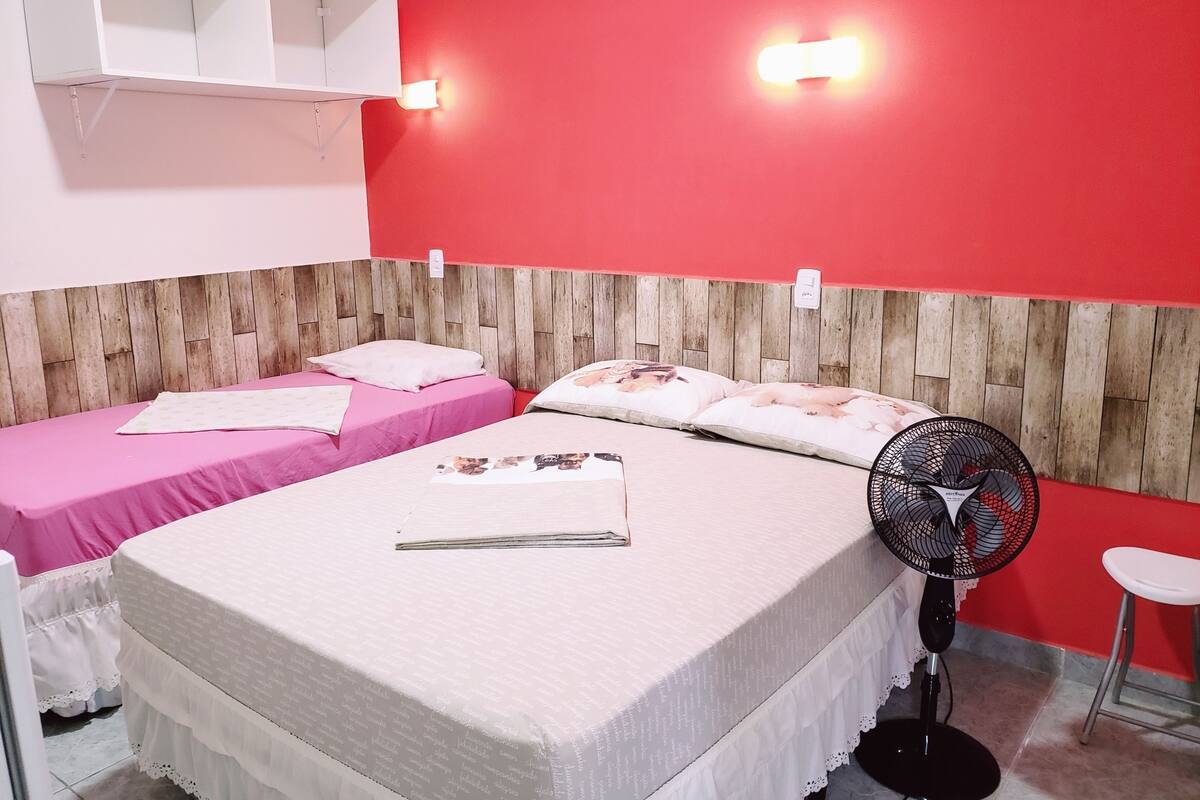 Ein Zimmer mit zwei Betten, einem hölzernen Kopfteil, einem an der Wand befestigten Regal, einem Standventilator und einem weißen Hocker.
