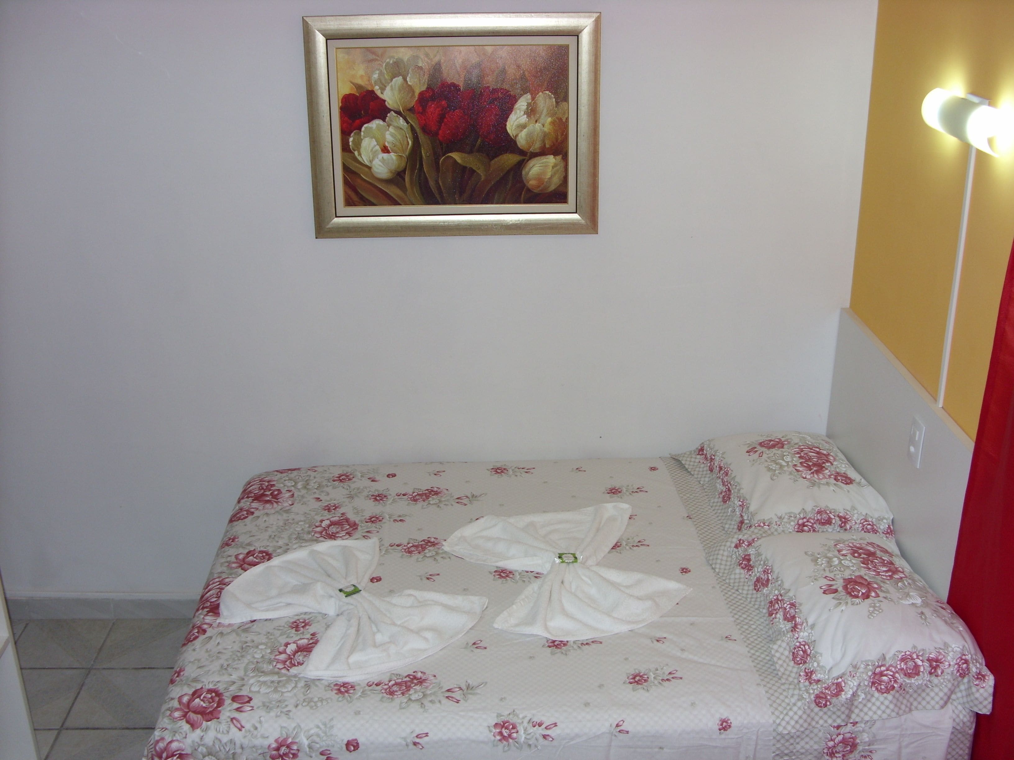 A bedroom with a floral bedspread, a painting of flowers on the wall, and a bedside lamp.