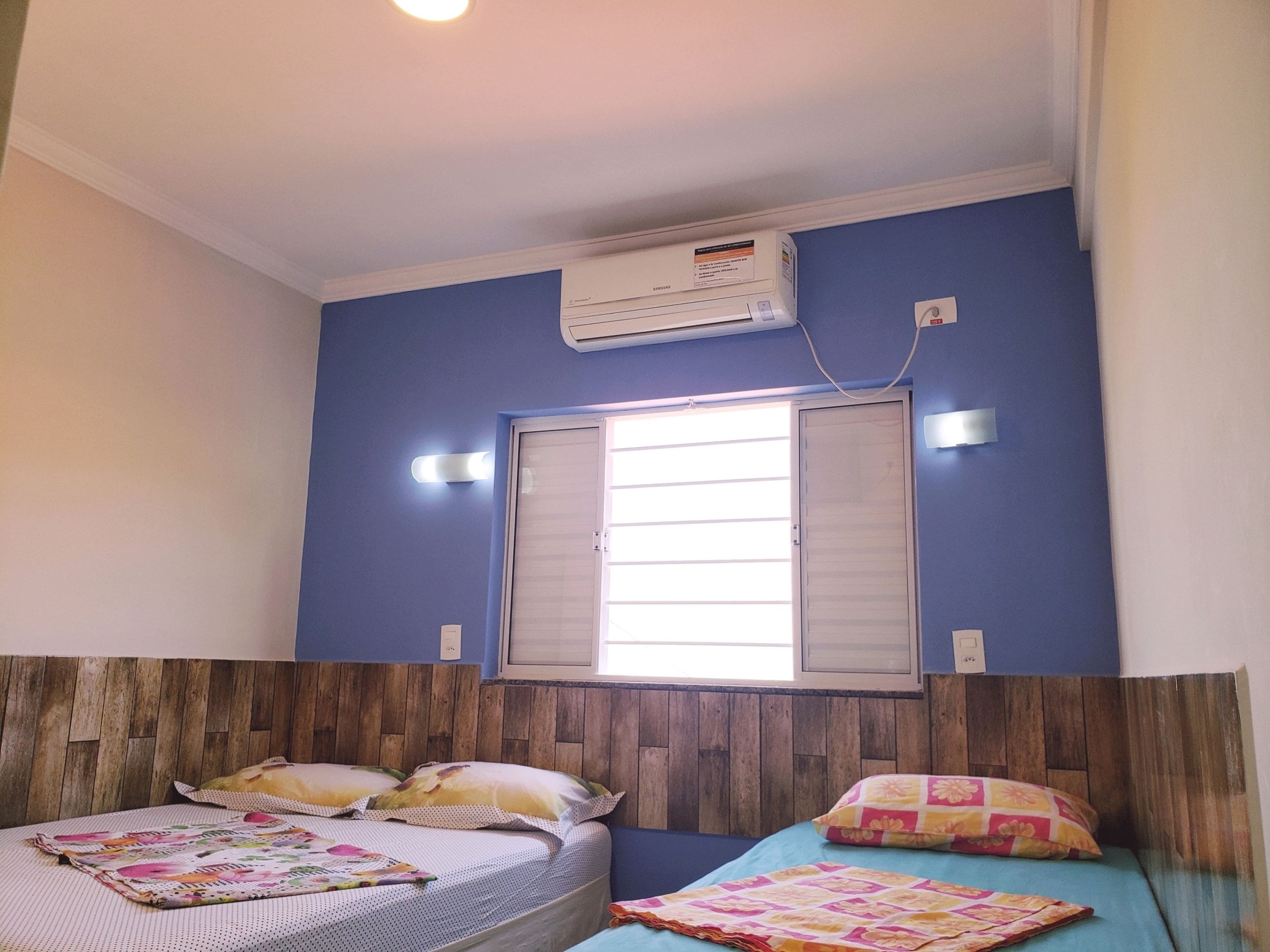 A room with two beds, a window with shutters, an air conditioner, and a wall-mounted light fixture.