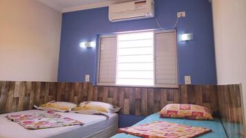 A room with two beds, a window with shutters, an air conditioner, and a wall-mounted light fixture.
