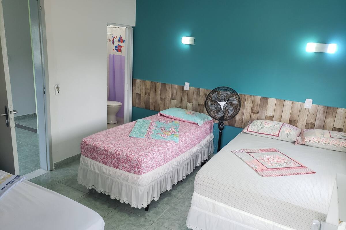 Ein Hotelzimmer mit zwei Betten, einem hölzernen Kopfteil, einem Standventilator und einem durch eine offene Tür einsehbaren Badezimmer.