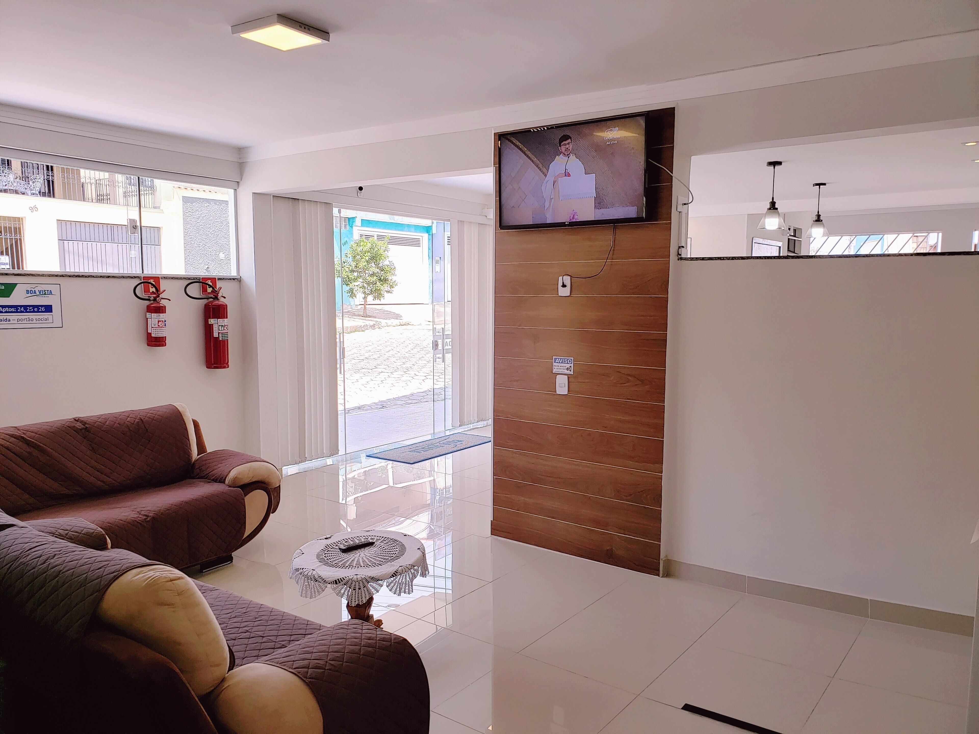 A modern reception area with a television, a wooden panel wall, a glass table with a remote, and a red fire extinguisher.