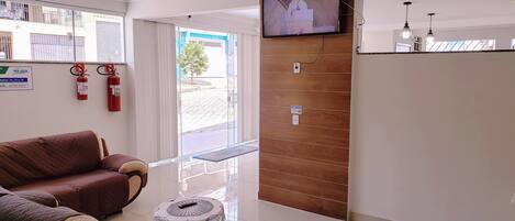 A modern reception area with a television, a wooden panel wall, a glass table with a remote, and a red fire extinguisher.
