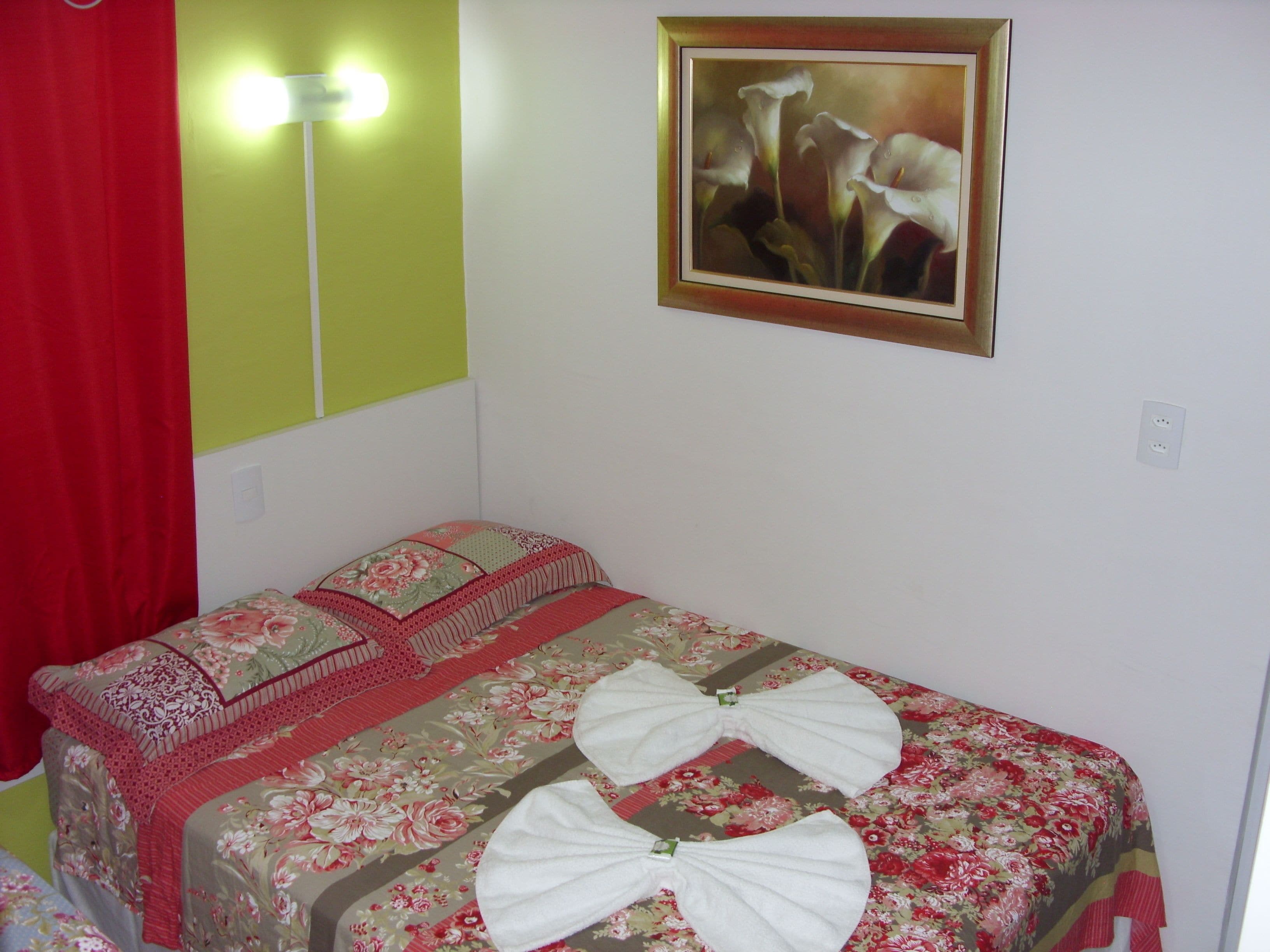Una cama pequeña y bien hecha con ropa de cama floral y dos toallas, una cortina roja, una lámpara fijada a la pared y un cuadro enmarcado con flores.