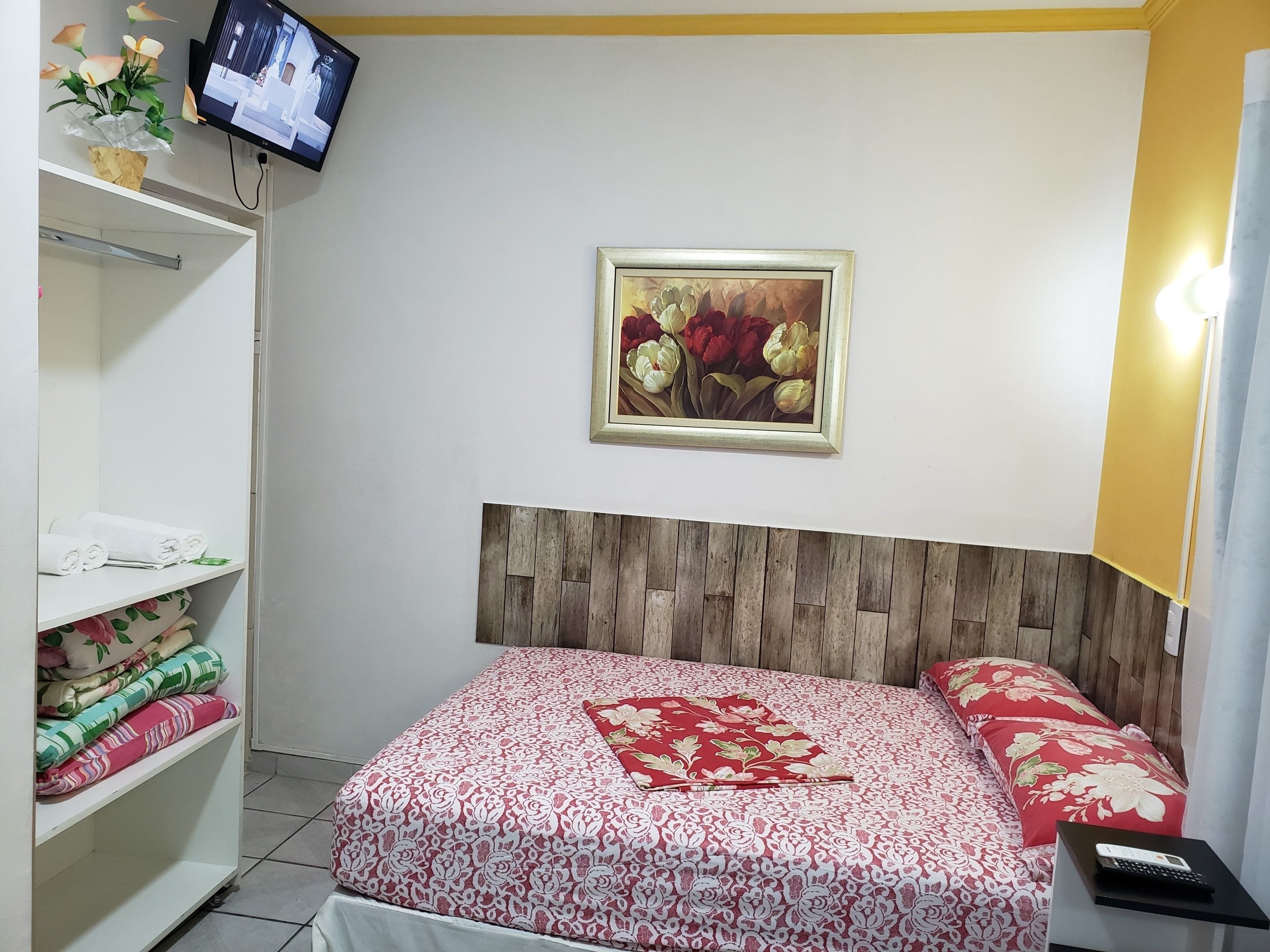 Ein ordentlich eingerichtetes Hotelzimmer mit einem hölzernen Kopfteil, einem Bett mit blumigem Bettzeug, einer Ablage mit gefalteten Handtüchern, einem gerahmten Blumenbild an der Wand und einem Fernseher, der über der Ablage angebracht ist.