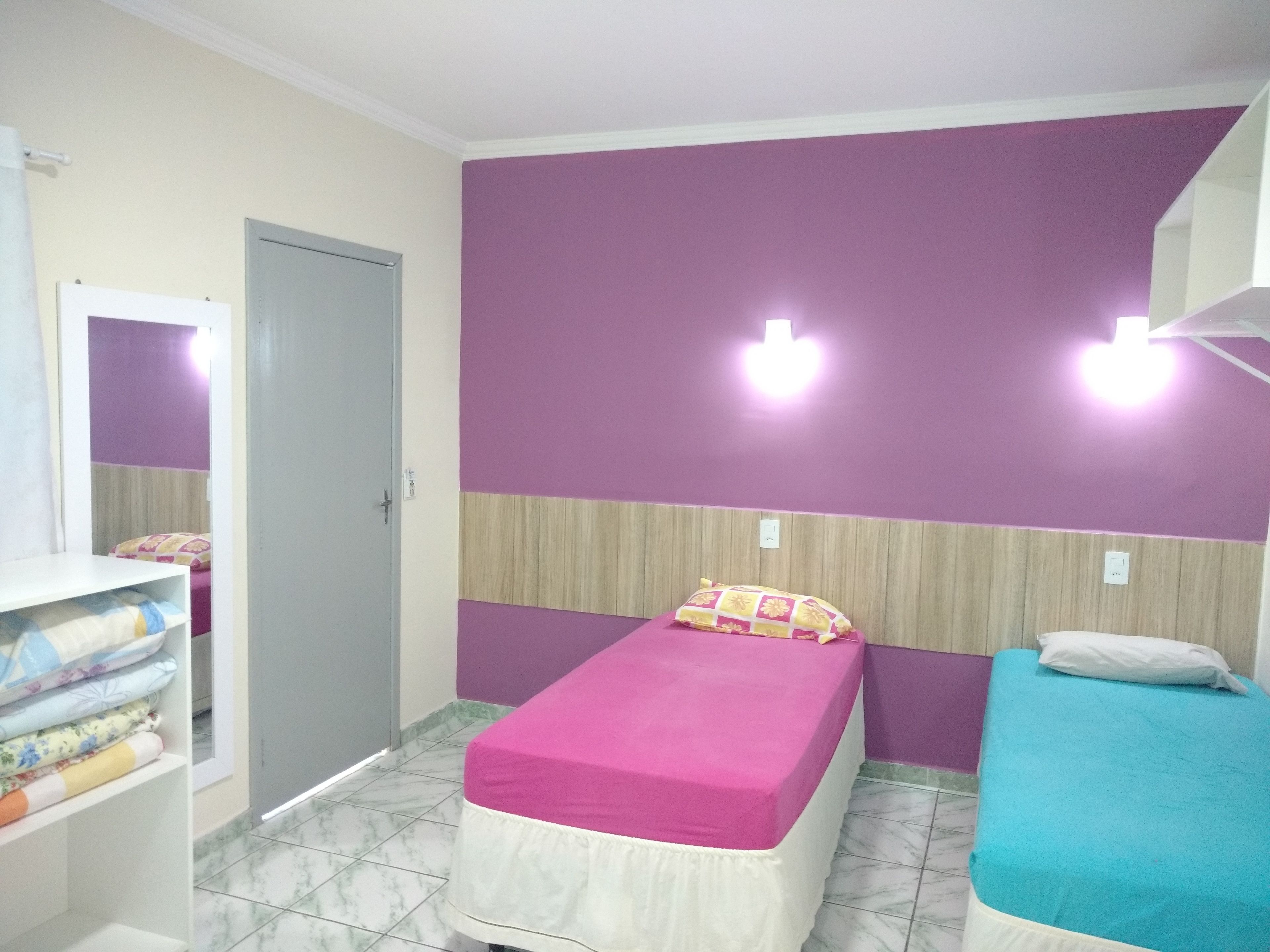 Une chambre avec deux lits, l’un recouvert d’une couvre-lit rose et l’autre d’une couvre-lit bleue. On y trouve une armoire contenant des vêtements pliés et un miroir accroché au mur.