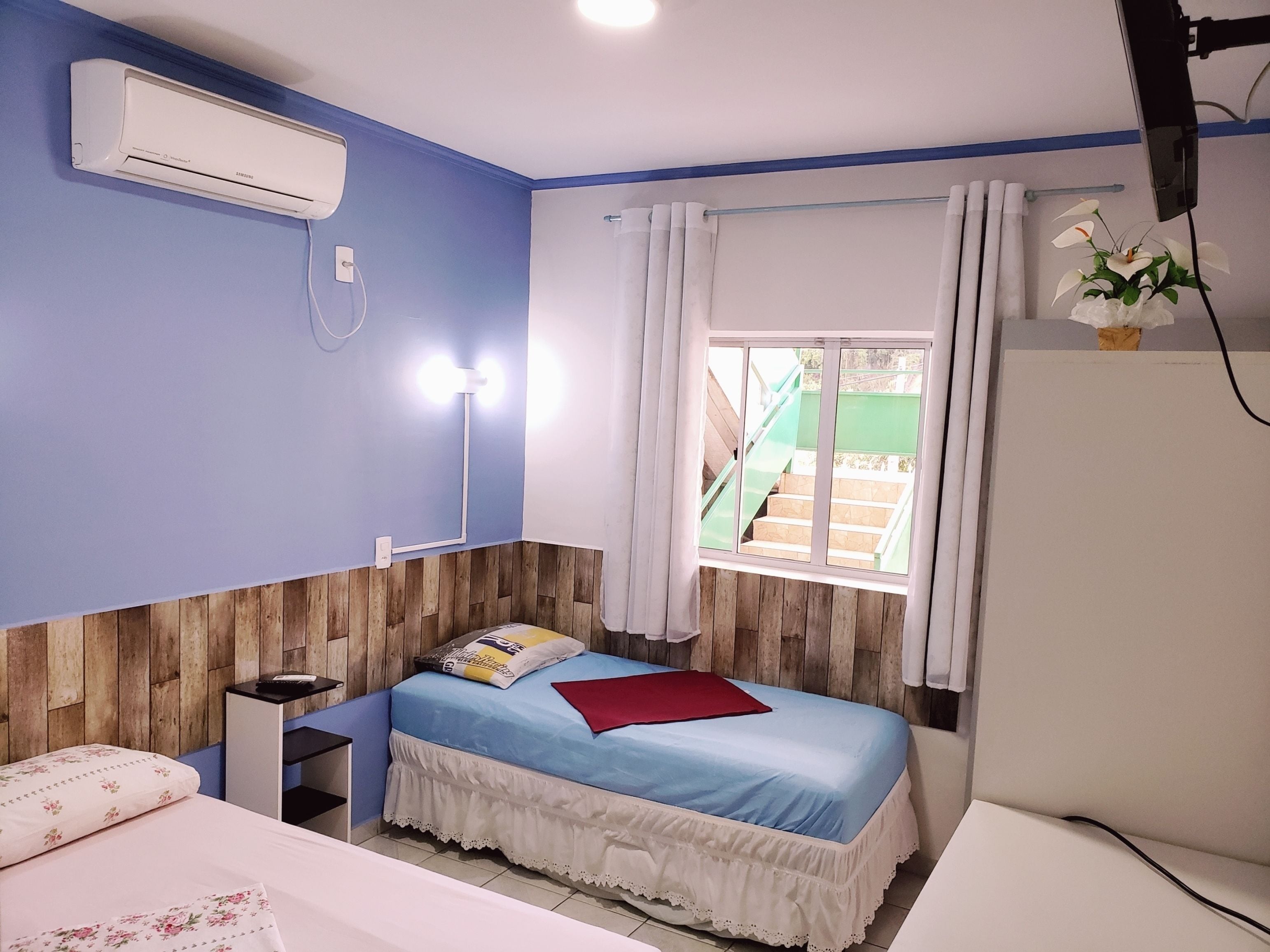 A bedroom with a bed, bedside table, air conditioning unit, window with curtains, and a view of stairs outside.