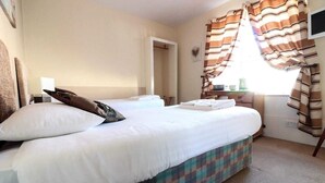 Een net opgemaakt bed met witte lakens en een geruit plaid, een nachtkastje met een lamp, en een raam met gestreepte gordijnen.