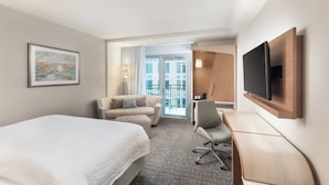 A hotel room with a bed, a desk, a chair, a TV, a couch, and a window with a view of buildings. - Courtyard by Marriott Baton Rouge Downtown (Baton Rouge)