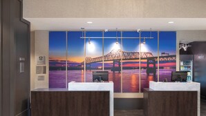 A hotel reception area with a large mural of a sunset over a bridge, two reception desks, a refrigerator stocked with beverages, and a sign indicating the availability of rooms. - Courtyard by Marriott Baton Rouge Downtown (Baton Rouge)