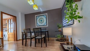 Een eetruimte met een tafel, stoelen, een aan de muur hangend schilderij, een potplant en een lamp.