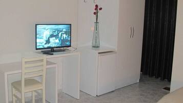 A small, clean room with a television on a white desk, a white chair, a white wardrobe, and a vase with flowers on top of the wardrobe.