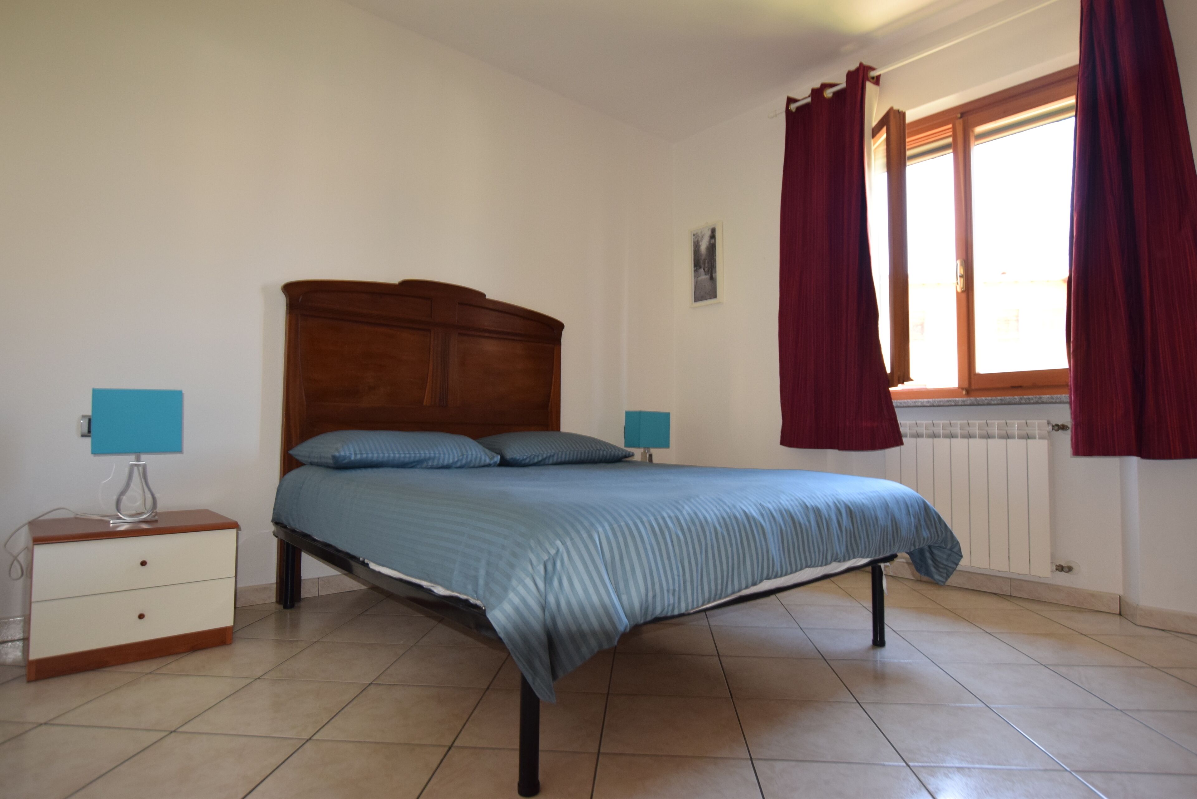 Une chambre à coucher avec un lit, deux tables de chevet, une fenêtre avec des rideaux et un radiateur.
