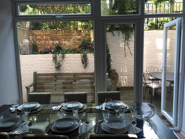 A dining area with a glass table set for six, surrounded by chairs, with a view of a brick wall and greenery outside.