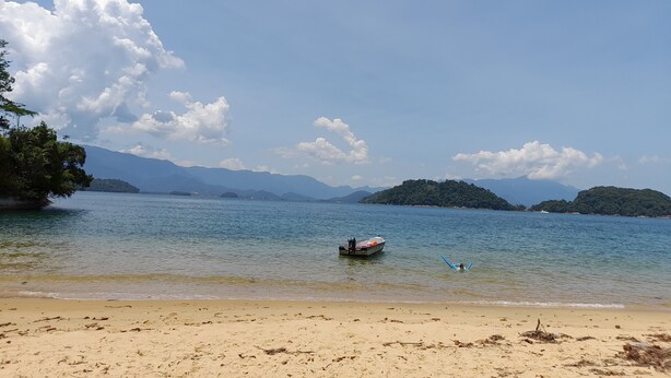 A beach with a boat anchored near the shore, surrounded by clear water and distant mountains. - Casa de Maria (Angra dos Reis)