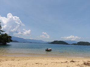 A beach with a boat anchored near the shore, surrounded by clear water and distant mountains. - Casa de Maria (Angra dos Reis)