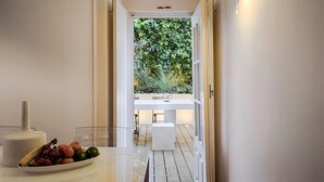 Un couloir étroit avec une table blanche et un saladier de fruits. Un verre d’eau est posé sur la table. Le couloir mène à un espace extérieur doté d’une terrasse en bois et offrant une vue sur la verdure.