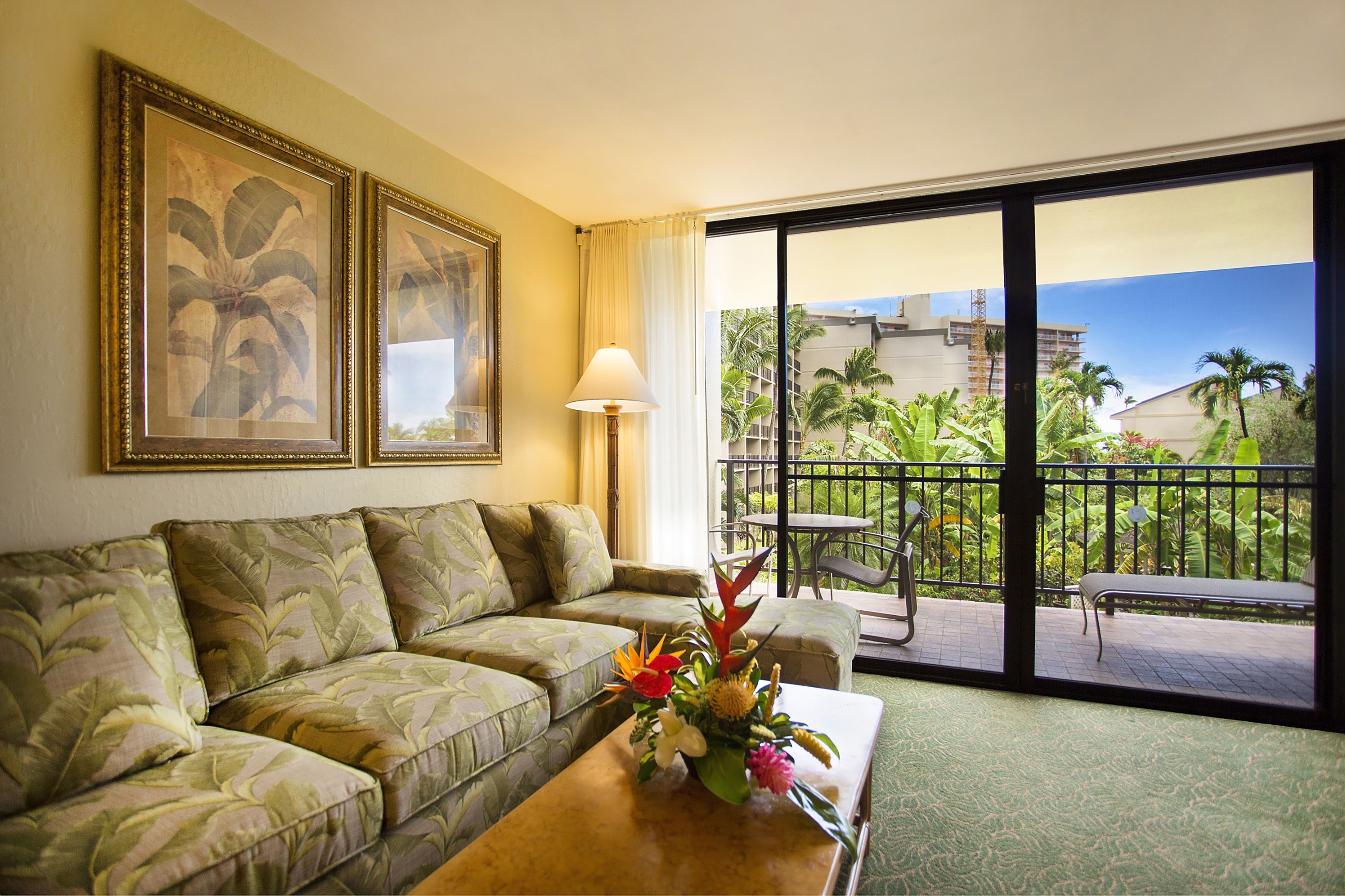 A hotel room with a sofa, a coffee table with a floral arrangement, a lamp, and a balcony with a view of palm trees and buildings.