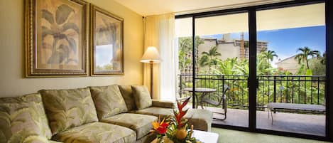 A hotel room with a sofa, a coffee table with a floral arrangement, a lamp, and a balcony with a view of palm trees and buildings.