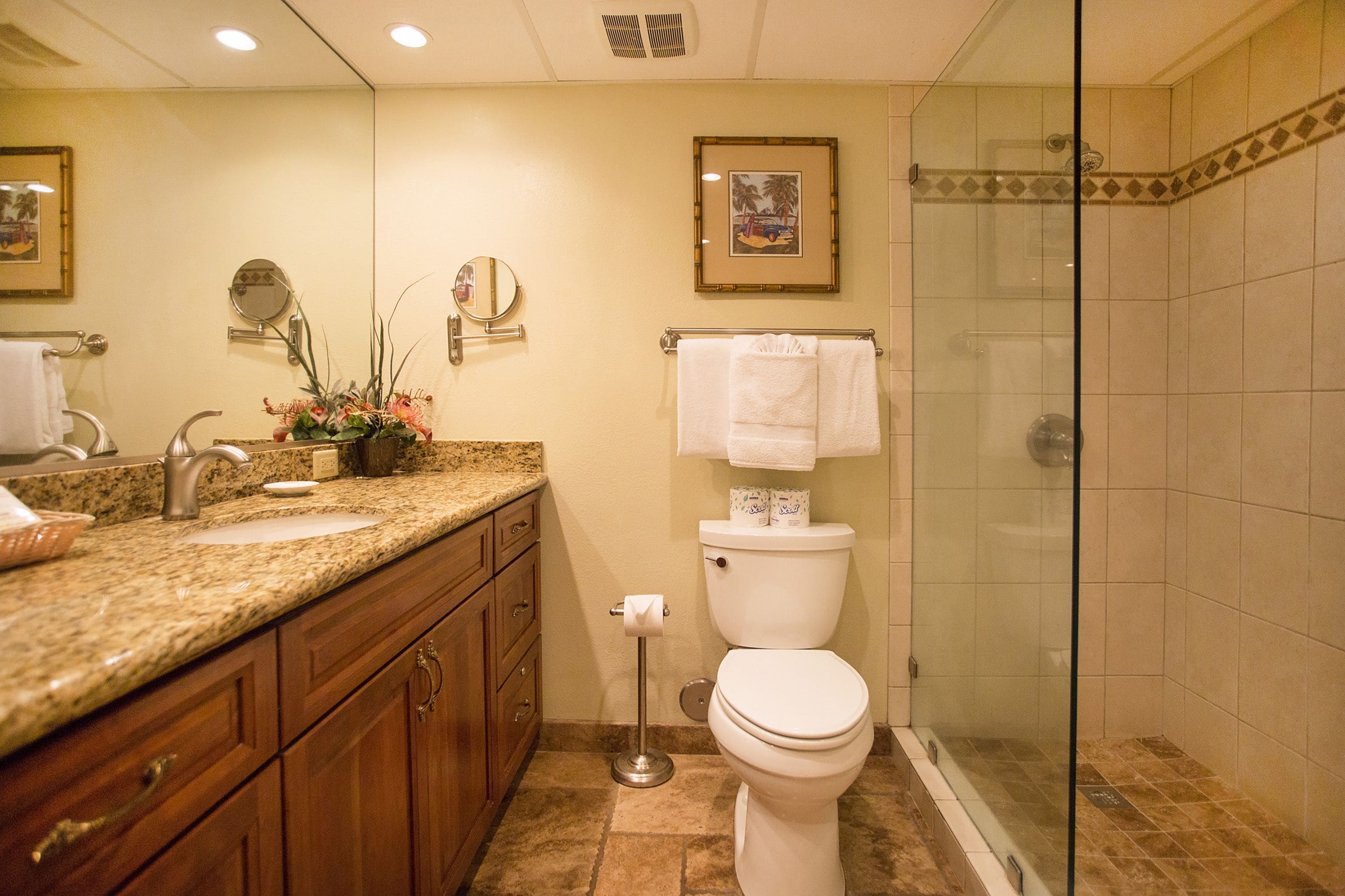 A bathroom with a granite countertop sink, wooden cabinets, a toilet, and a glass-enclosed shower.