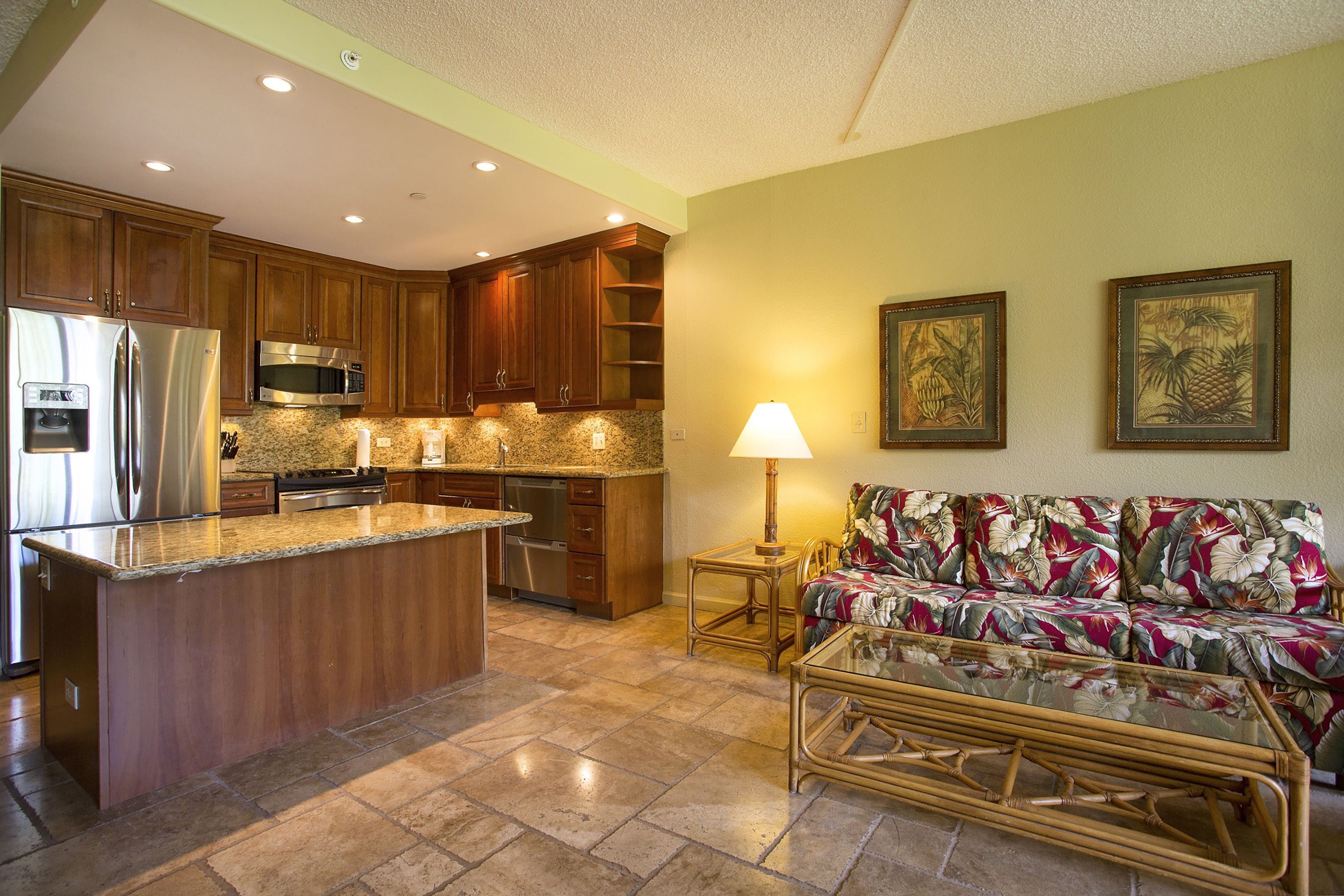 A kitchen with wooden cabinets, stainless steel appliances, and a central island. There is a dining area with a glass-top table and a sofa with a floral pattern.